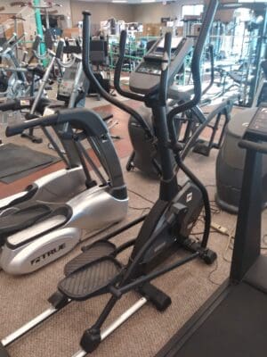 A black elliptical machine with a digital console stands in a gym among treadmills and stationary bikes.