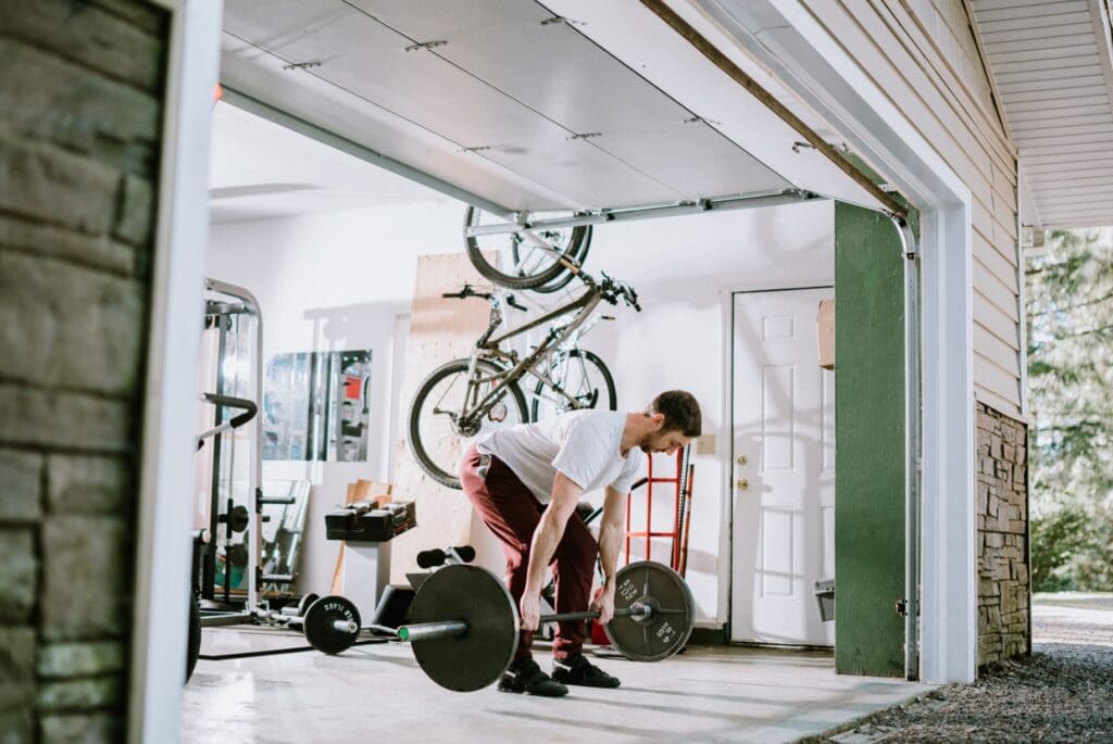 A man in a white t-shirt and red pants deadlifts a barbell in a well-organized garage gym with various equipment and bikes.