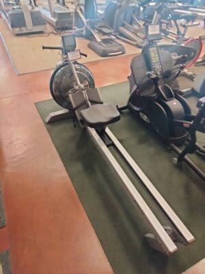 Rowing machine with a metal frame and seat, placed on a green mat in a gym alongside other cardio equipment.