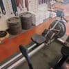 A rowing machine with a black seat, foot straps, and a flywheel sits on a green mat in a gym beside other cardio equipment.