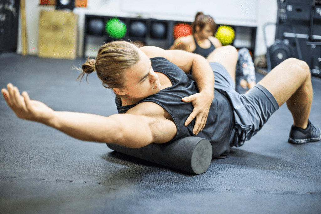 A man in a black tank top and shorts uses a foam roller for muscle recovery, stretching his arm while lying on the gym floor.