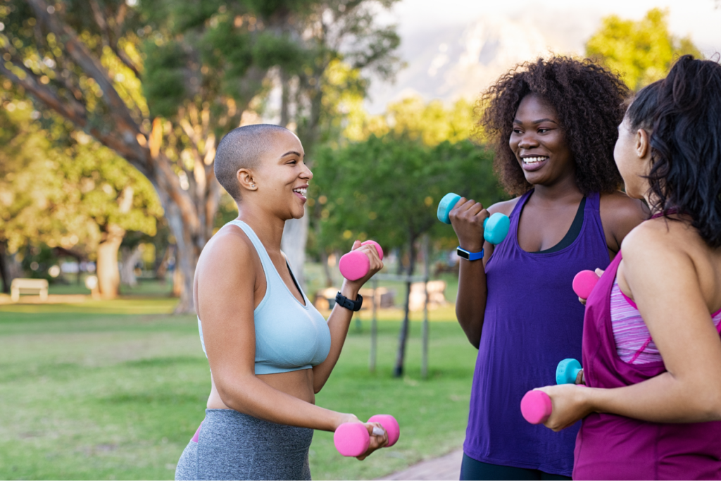 Three women in athletic wear smile while lifting dumbbells together in a sunny park with trees and mountains behind them.