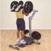 A man in a yellow tank top and shorts performs a squat on a plate-loaded machine, pushing upward with weighted resistance.