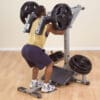 A man in a yellow tank top and shorts performs a squat on a plate-loaded machine, pushing upward with weighted resistance.
