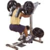 A man in a yellow tank top and shorts performs a squat on a plate-loaded machine, pushing upward with weighted resistance.