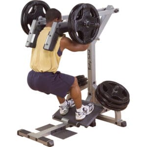 A man in a yellow tank top and shorts performs a squat on a plate-loaded machine, pushing upward with weighted resistance.