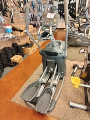 A gray elliptical machine with moving handlebars and foot pedals sits in a gym, surrounded by fitness equipment and weights.