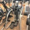 A gray elliptical with a digital console, moving handlebars, and foot pedals stands in a gym among other fitness equipment.