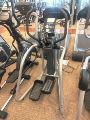 A gray elliptical with a digital console, moving handlebars, and foot pedals stands in a gym among other fitness equipment.