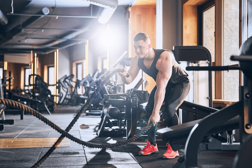 Man in a gym performing battle rope exercises, surrounded by fitness equipment and lit by natural sunlight through windows.