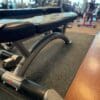 A black flat weight bench with a silver frame sits on a rubber gym floor, with exercise equipment visible in the background.