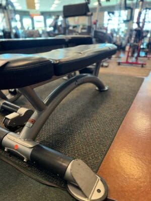 A black flat weight bench with a silver frame sits on a rubber gym floor, with exercise equipment visible in the background.
