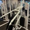 A white incline press machine with padded seating and weights stands in a gym, surrounded by strength training equipment.