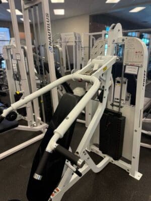 A white incline press machine with padded seating and weights stands in a gym, surrounded by strength training equipment.