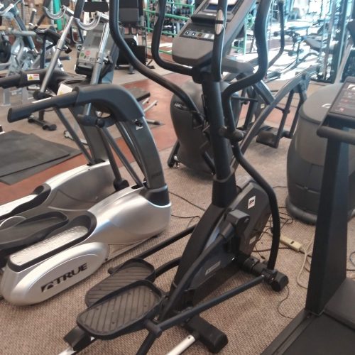 A black elliptical machine with a digital console stands in a gym among treadmills and stationary bikes.