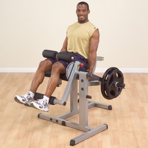 A man in a yellow tank top and shorts uses a seated leg extension machine with weights in a gym with a light wood floor.