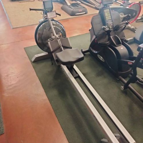 Rowing machine with a metal frame and seat, placed on a green mat in a gym alongside other cardio equipment.